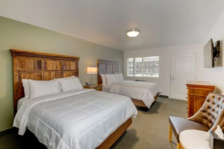 A hotel room with two queen beds, a wooden headboard, and a flat-screen TV on the wall.