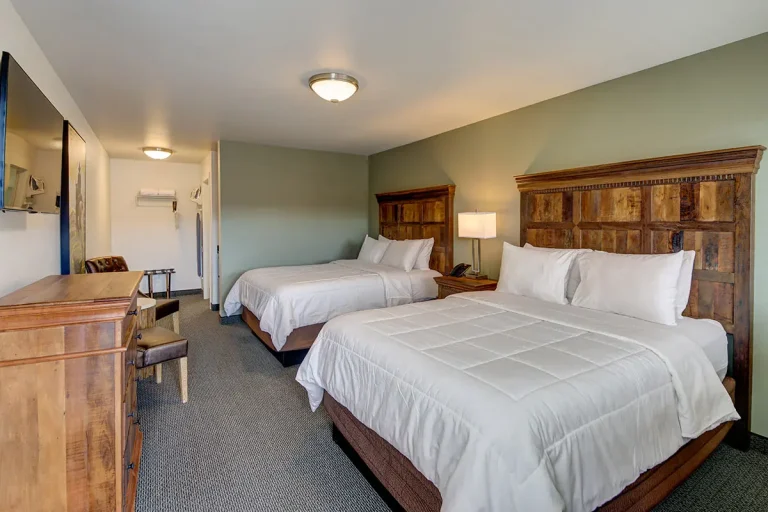 A hotel room with two queen beds, wooden headboards, a flat-screen TV, a desk, and chairs.