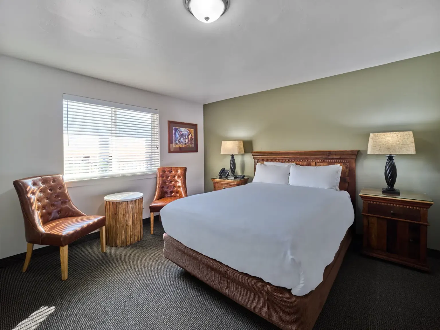 A hotel room with a queen bed, two nightstands, two leather chairs, and a window with blinds.
